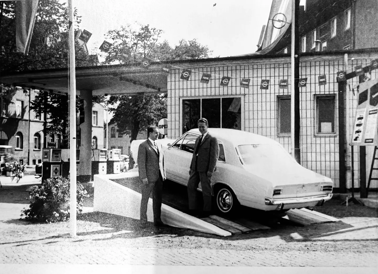 Historische Aufnahme: Opel Rekord und zwei Verkäufer vor der Tankstelle mit Opel-Fahnen